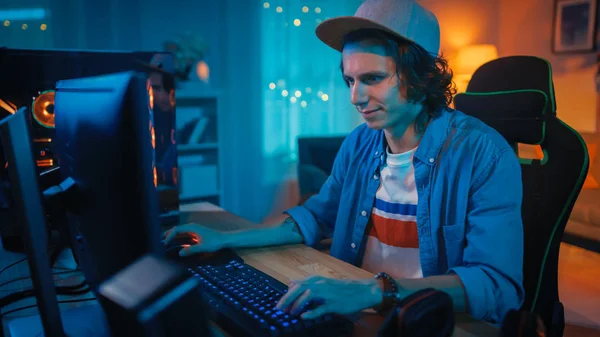 Jogador animado em Cap Jogando videogame online em seu computador pessoal. O quarto e o PC têm luzes conduzidas quentes coloridas do néon. O Young Man está a usar um boné. Noite aconchegante em casa . — Fotografia de Stock