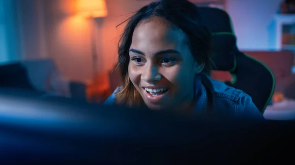 Linda surpreendido e animado jovem preto menina blogueiro assistindo vídeo de ação em um computador. Ela tem cabelo escuro e olhos castanhos. Screen Adiciona Reflexões ao Seu Rosto. Quarto acolhedor é lit com luz quente . — Fotografia de Stock