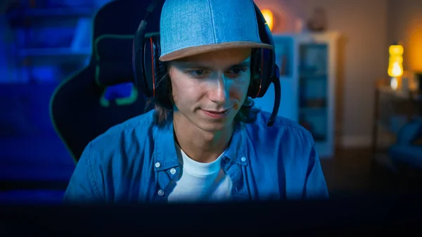 Populäre Gamer spielt Online-Videospiel auf seinem PC. Zimmer und PC haben bunte warme Neon-LED-Lichter. Der junge Mann trägt Kopfhörer mit Mikrofon. Gemütlicher Abend zu Hause. — Stockfoto