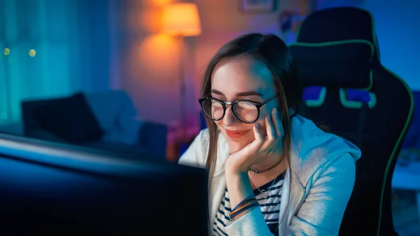 Mooi gelukkig en sentimenteel jong meisje blogger kijken naar Video's op een computer. Ze heeft donker haar en draagt een bril. Scherm voegt reflecties toe aan haar gezicht. Gezellige kamer verlicht met warm licht. — Stockfoto