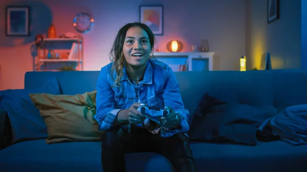 Beautiful Excited Young Black Gamer Girl Sitting on a Couch and Playing Video Games on a Console. She Plays with a Wireless Controller. Cozy Room is Lit with Warm and Neon Light.