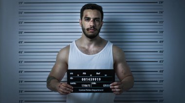 Bir Polis Karakolu'nda Tutuklanan Dayak Adam Front View Mugshot için Pozlar. O Singlet giyer, Ağır Çürük ve Placard tutar. Arka Planda Yükseklik Grafiği. Koyu Soğuk Işıklar, Vignette Filtre ile Shot