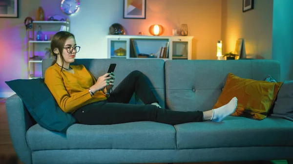 Όμορφη ενθουσιασμένος νεαρό κορίτσι σε γυαλιά κάθεται σε έναν καναπέ και χρησιμοποιώντας το smartphone της. Χαμογελάει ενώ η τηλεόραση παίζει στο παρασκήνιο. Η οθόνη προσθέτει αντανακλάσεις στο πρόσωπό της. Το Cozy Room είναι αναμμένο με θερμό φως. — Φωτογραφία Αρχείου