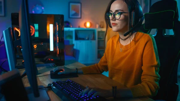 Fille joueuse excitée et concentrée dans des lunettes et un casque avec un micro jouant jeu vidéo de stratégie en ligne sur son ordinateur personnel. Chambre et PC ont des lumières LED au néon chaud coloré. Soirée confortable à la maison . — Photo