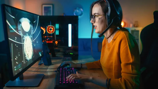 Vzrušená Gamer Girl v náhlavní soupravě s mikrofonem přehráváním online videohry na jejím osobním počítači. Mluví s ostatními hráči. V místnosti a počítači jsou barevné teplé neonové indikátory LED. Útulný večer doma. — Stock fotografie