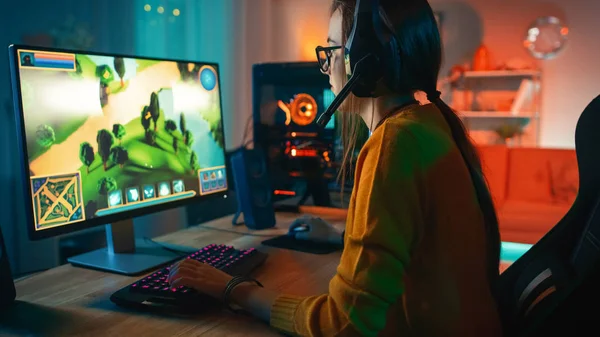 Fille joueuse excitée et concentrée dans des lunettes et un casque avec un micro jouant jeu vidéo de stratégie en ligne sur son ordinateur personnel. Chambre et PC ont des lumières LED au néon chaud coloré. Soirée confortable à la maison . — Photo