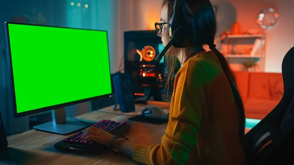 Возбужденная и сконцентрированная девушка-геймер в очках и наушниках с микрофоном, играющим на её персональном компьютере с шаблоном макета зелёного экрана. Красочные теплые неоновые огни. Уютный вечер дома . — стоковое фото