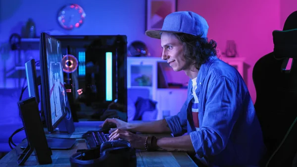 Jugador profesional que juega el videojuego en línea del tirador en primera persona en su computadora personal potente con luces led de neón coloridas. El joven lleva una gorra. Sala de estar Lit con luz de neom rosa . —  Fotos de Stock