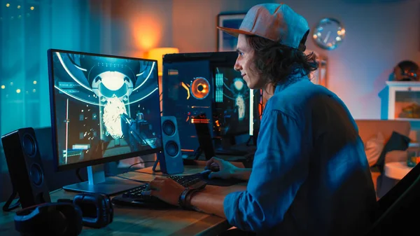 专业玩家玩第一人称射击在线视频游戏在他的强大的个人计算机与五颜六色的霓虹灯。年轻人戴着帽子。客厅用暖灯照明。晚上. — 图库照片