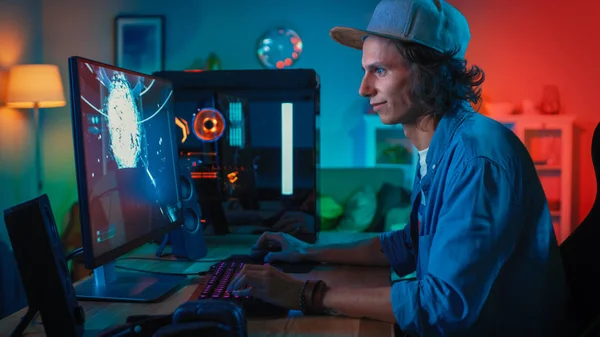 专业玩家玩第一人称射击在线视频游戏在他的强大的个人计算机与五颜六色的霓虹灯。年轻人戴着帽子。客厅用暖灯照明。晚上. — 图库照片