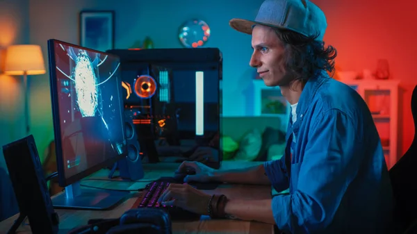 Jugador profesional que juega el videojuego en línea del tirador en primera persona en su computadora personal potente con luces led de neón coloridas. El joven lleva una gorra. Sala de estar Lit con lámparas calientes. Buenas noches. . —  Fotos de Stock