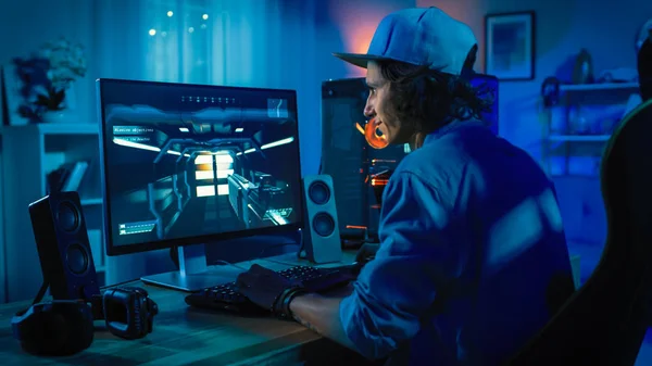 Professionelle Gamer, die Ego-Shooter-Online-Videospiel auf seinem leistungsstarken Personal Computer mit bunten Neonröhren spielen. Der junge Mann trägt eine Mütze. Wohnzimmer beleuchtet in Low-Key-Stil. Abend. — Stockfoto