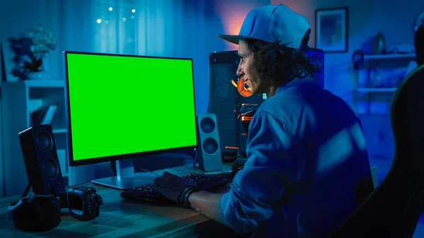 Joueur professionnel jouant au jeu vidéo en ligne sur son ordinateur personnel puissant avec des lumières LED au néon coloré. Maquette d'écran vert. Le jeune homme porte une casquette. Salon avec lampes chaudes. Bonsoir. . — Photo