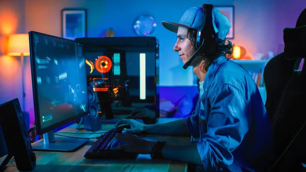 Profesjonalny Gamer gra strzelanka z perspektywy pierwszej osoby online gry wideo na jego potężny komputer osobisty. Pokój i komputer mają kolorowe Neon LED Lights. Młody człowiek nosi czapkę. Przytulny wieczór w domu. — Zdjęcie stockowe