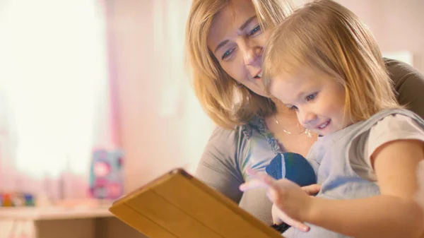 Mooie moeder en haar schattige kleine dochter zittend op de vloer en het lezen van kinderboeken op een tablet computer. Zonnige woonkamer. — Stockfoto