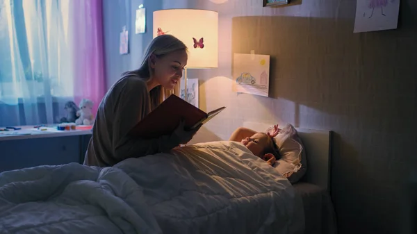 Joven madre cariñosa lee cuentos para dormir a su pequeña hija hermosa que se va a dormir en su cama . — Foto de Stock