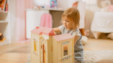 Çocuk Odasında: Sevimli Mutlu Küçük Kız Doll House ile oynamak ve bir halı üzerinde oturan. Eğlenen Çocuklu Güneşli Oturma Odası.