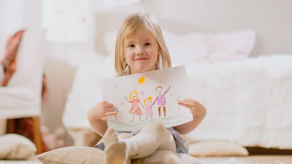 Söt ung leende flicka sitter på kuddar och visar ritning av hennes familj. Soligt vardagsrum. — Stockfoto