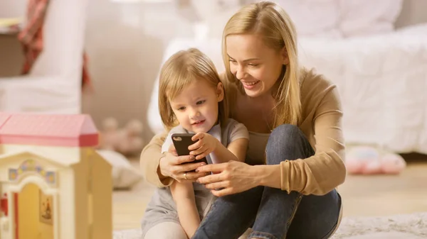 Mooie moeder en haar kleine dochter hebben goede tijd praten, moeder houdt smartphone. Zonnige kinderkamer vol speelgoed. — Stockfoto