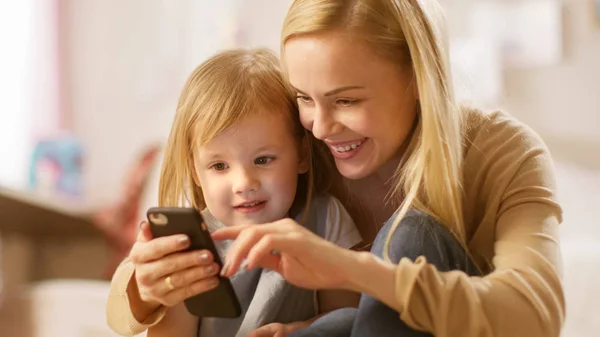 Close-up Tiro de uma bela jovem mãe com sua filhinha brincando com Smartphone. A mãe mostra-lhe algo interessante. Quarto ensolarado para crianças . — Fotografia de Stock
