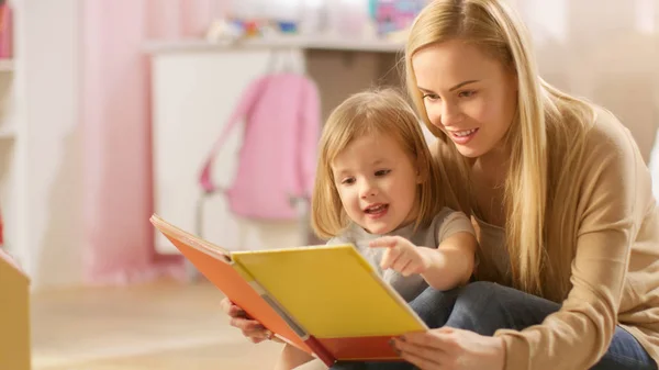 Mooie jonge moeder en haar schattige kleine dochter lezen kinderboek samen. Kinderkamer is roze en volledige van speelgoed. — Stockfoto