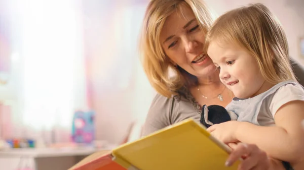 Close-up Shot of a Beautiful Young Mother and Her Cute Little Daughter Read Children 's Book Together. Habitación de los niños es de color rosa y lleno de juguetes . — Foto de Stock