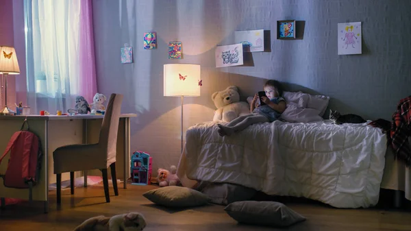 Linda niña se encuentra en su cama y mira dibujos animados en un teléfono inteligente. Su gatita está a su lado tocando. Su lámpara de pie está encendida . — Foto de Stock