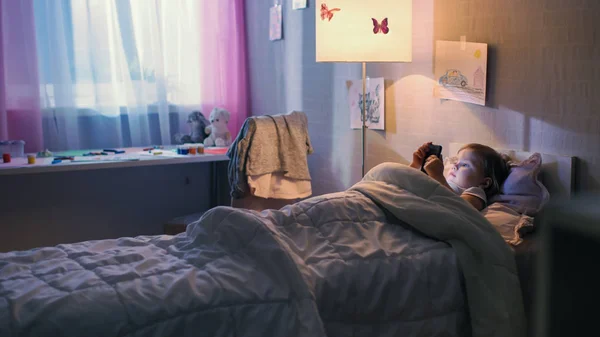 Το χαριτωμένο μικρό κορίτσι βρίσκεται στο κρεβάτι και κινούμενα σχέδια Ρολόγια σε Smartphone. Το Επιδαπέδιο φωτιστικό αναμμένο. — Φωτογραφία Αρχείου