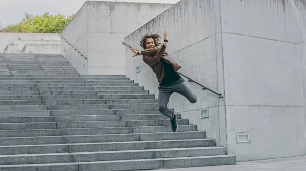 Jovem alegre e feliz com salto de cabelo longo enquanto caminha pelas escadas. Ele está a usar um casaco de couro castanho. Cena filmada em um parque de concreto urbano. Dia é brilhante . — Fotografia de Stock