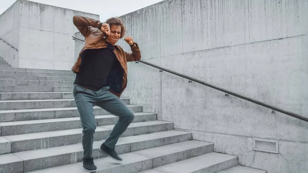 Joven alegre y feliz con el pelo largo bailando activamente mientras camina por las escaleras. Lleva una chaqueta de cuero marrón. Escena filmada en un parque urbano de hormigón junto al centro de negocios. Soleado. . — Foto de Stock