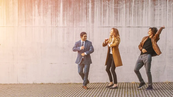 Den glade jenta og den glade unge mannen med langt hår danser aktivt på en gate. Forretningsmann i dress slutter seg til dem. De har på seg brun skinnjakke og jakke. Solskinnsdag . – stockfoto