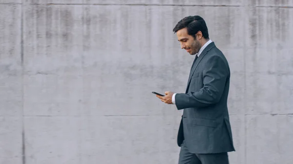 Happy Businessman está caminhando com um smartphone em uma rua ao lado de uma parede de concreto urbano. Ele está a usar um fato cinzento. Dia de sol . — Fotografia de Stock