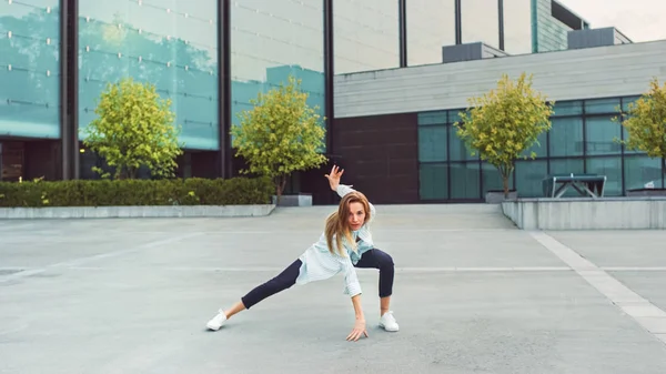 Mujer joven feliz bailando activamente en una calle. Lleva camisa blanca y pantalones vaqueros azules. Escena filmada en un parque urbano de hormigón junto al centro de negocios . — Foto de Stock