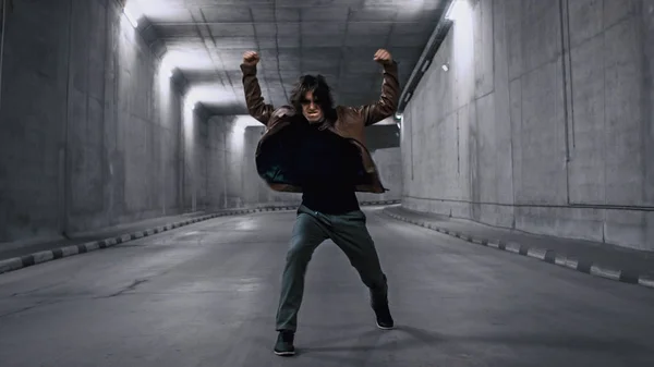 Серйозний прохолодний молодий чоловік хіпстера з довгим волоссям енергійно танцює хіп-хоп в освітленому бетонному тунелі. Він носить коричневу шкіряну куртку. — стокове фото