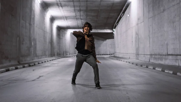 Serious Cool Young Hipster Man con i capelli lunghi è Energeticamente Dancing Hip Hop in un tunnel di cemento Lit. Indossa una giacca di pelle marrone. . — Foto Stock