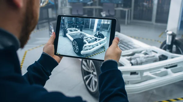 Ingeniero con gafas y barba escanea el prototipo de chasis de coche eléctrico con ruedas, baterías y motor con un software de realidad aumentada en una tableta en un laboratorio de desarrollo de alta tecnología . —  Fotos de Stock