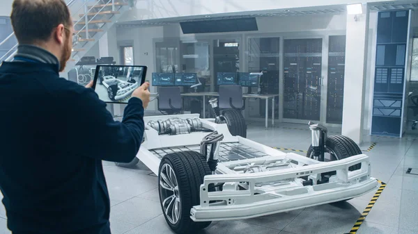 Gözlük ve Sakallı Mühendis, Yüksek Teknoloji Geliştirme Laboratuvarında Tablet Bilgisayarda Artırılmış Gerçeklik Yazılımı ile Tekerlekler, Piller ve Motor ile Elektrikli Araba Şasi Prototipini Tarıyor.