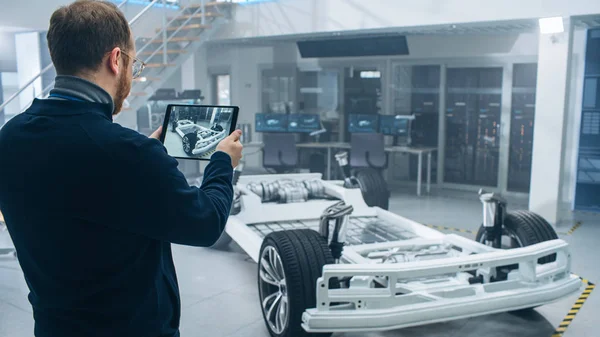 Manlig ingenjör arbetar i en hög teknisk utveckling anläggning som innehar en Tablet PC. Han står bredvid en elektrisk bil chassi prototyp med batterier och hjul. — Stockfoto