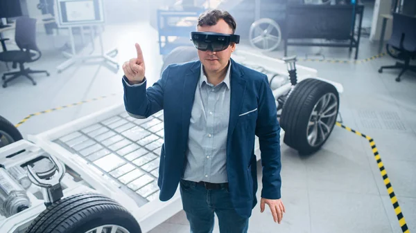 Ingénieur automobile utilisant casque de réalité augmentée faisant des gestes touchants. Dans le concept d'installation de laboratoire d'innovation, le cadre du véhicule comprend les roues, la suspension, le moteur et la batterie . — Photo