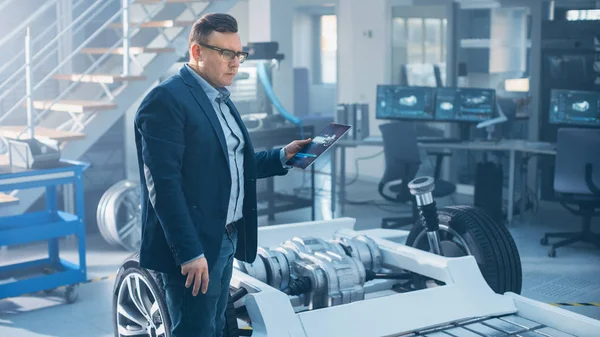 Ingénieur avec lunettes Fonctionne sur une tablette à côté d'un prototype de châssis de voiture électrique avec roues, batteries et moteur dans un laboratoire de développement de haute technologie . — Photo