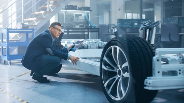 Ingénieur avec lunettes Fonctionne sur une tablette à côté d'un prototype de châssis de voiture électrique avec roues, batteries et moteur dans un laboratoire de développement de haute technologie . — Photo