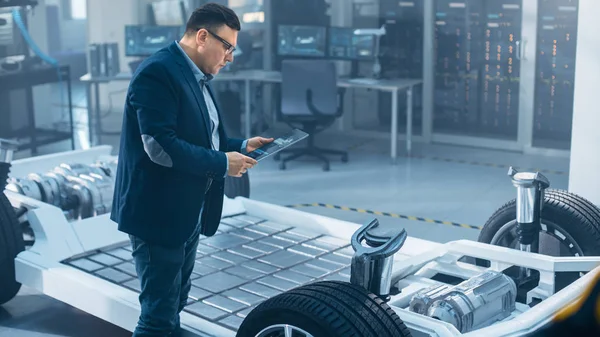 Ingeniero de Diseño Automotriz Viendo el Dibujo Técnico de un Prototipo de Chasis de Coche Eléctrico en una Tableta. Marco del vehículo del concepto de la instalación del laboratorio de la innovación incluye ruedas, motor y batería . —  Fotos de Stock