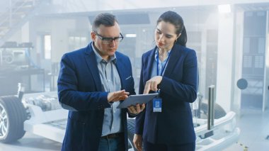 Tablet Bilgisayar Tutan Bir Yüksek Teknoloji Geliştirme Tesisinde Kadın ve Erkek Mühendis Çalışması. Onlar Piller ve Tekerlekler ile Elektrikli Otomobil Şasi Prototip Yanında Stand.