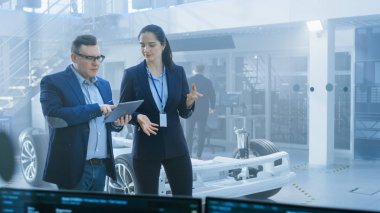 Kadın ve Erkek Mühendis Bir Tablet Bilgisayar Tutan Bir Yüksek Teknoloji Geliştirme Tesisi Walk ve Piller ve Tekerlekler ile Elektrikli Araba Şasi Prototip Pass.