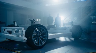 Otomotiv İnovasyon Tesisinde İki Otomobil Tasarım Mühendisinin Siluetleri. Onlar Tekerlekler, Süspansiyon, Hibrid Motor ve Pil içeren Elektrikli Otomobil Platformu Şasi Prototip üzerinde çalışıyoruz.