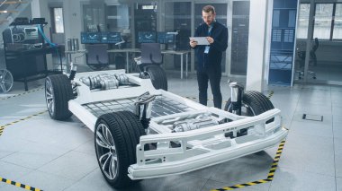 Gözlük ve Sakallı Mühendis, Yüksek Teknoloji Geliştirme Laboratuvarında Tablet Bilgisayarda Artırılmış Gerçeklik Yazılımı ile Tekerlekler, Piller ve Motor ile Elektrikli Araba Şasi Prototipini Tarıyor.