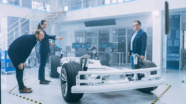 Engenheiros de design automotivo conversando enquanto trabalhava no protótipo de chassi de carro elétrico. Na estrutura do veículo do conceito da facilidade do laboratório da inovação inclui rodas, suspensão, motor e bateria . — Fotografia de Stock