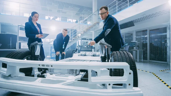 Ingenieros de diseño automotriz que hablan mientras trabajan en el prototipo del chasis del coche eléctrico. Marco del vehículo del concepto de la instalación del laboratorio de la innovación incluye ruedas, suspensión, motor y batería . —  Fotos de Stock