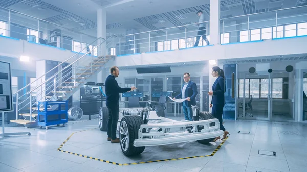 Automotive Design Engineers Talking työskennellessään sähköauton alustan prototyyppi. Innovaatiolaboratorio laitos käsite ajoneuvon runko sisältää pyörät, jousitus, moottori ja akku . — kuvapankkivalokuva