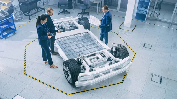 Automotive Design Engineers Talking while Working on Electric Car Chassis Prototype. In Innovation Laboratory Facility Concept Vehicle Frame Includes Wheels, Suspension, Engine and Battery. — Stock Photo, Image